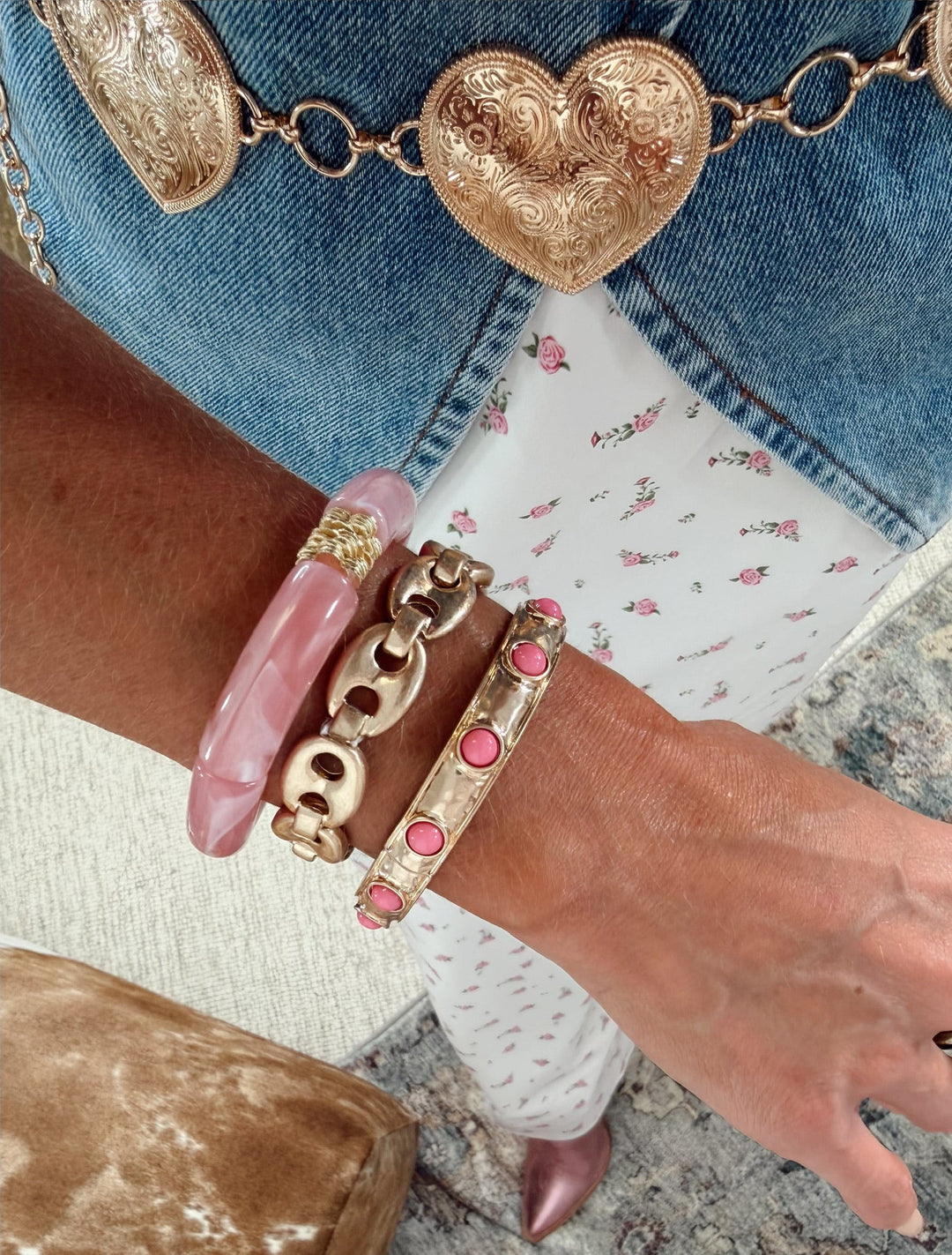 A bracelet stack  including a pink stone-embellished gold bangle, a chunky gold chain bracelet, and a translucent pink bangle, paired with blue jeans and a patterned top.